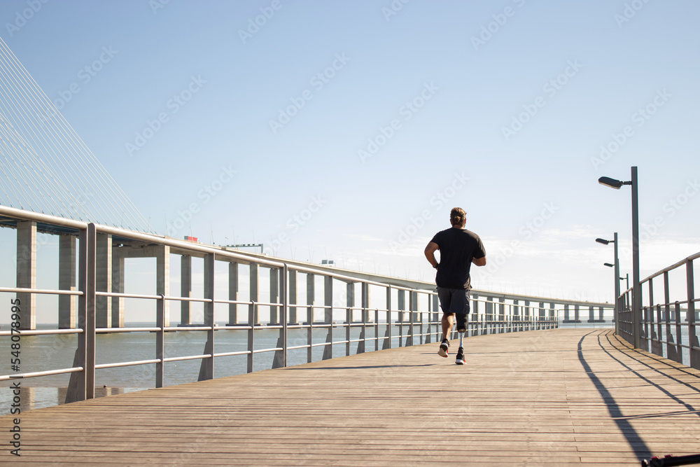 Back view of man with artificial leg jogging on bridge. Sporty man in casual clothes training outdoors in morning running on bridge over sea. Health care, active life of people with disability concept