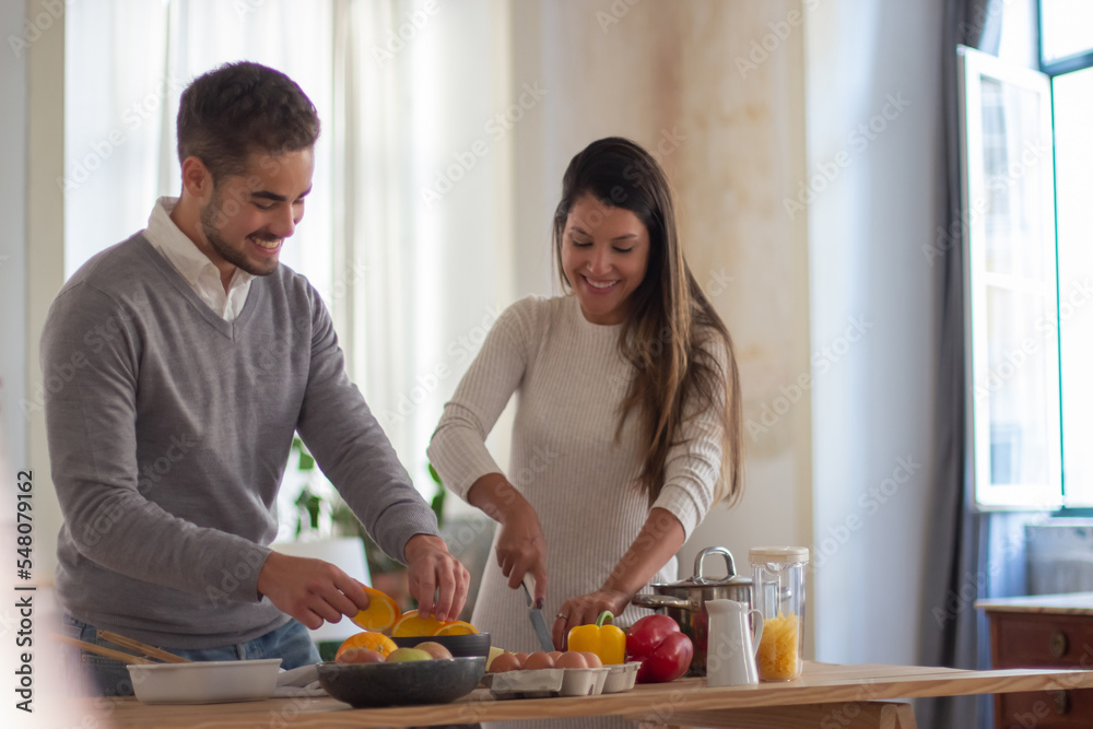Happy young couple cooking delicious salad together. Beautiful woman concentrated on cutting fresh fruit and vegetables and man helping her. Cooking food and healthy lifestyle concept