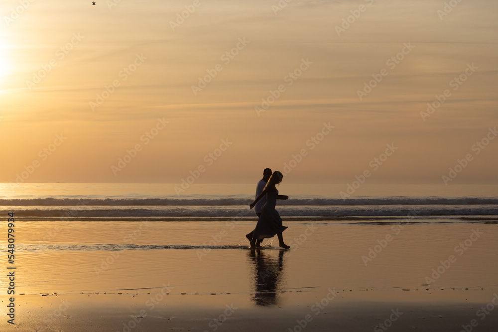 Naklejka premium Romantic couple walking on beach at sunset. Man and woman in casual clothes strolling along water at dusk. Love, family, nature concept