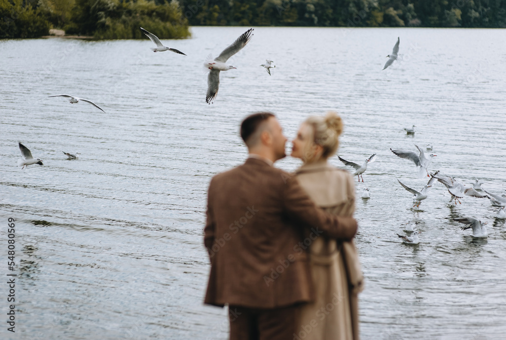 Φωτογραφία A stylish groom in a brown suit and a beautiful blonde model bride gently embrace, standing near the sea, a lake against the background of water with seagulls