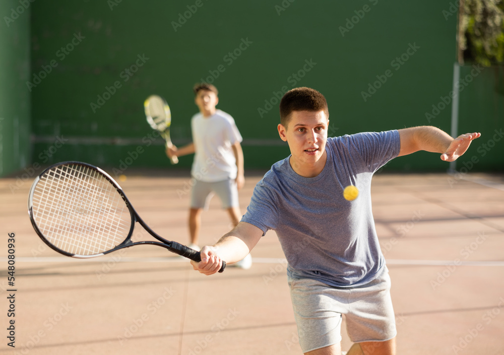 Young male pelota player hitting ball with racket during training game on outdoor Basque pelota fronton.