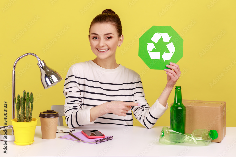 Delighted woman office manager holding recycling sign and pointing at plastic, glass bottles and cardboard package, thinking green. Indoor studio studio shot isolated on yellow background. Delighted woman office manager holding recycling sign and pointing at plastic, glass bottles and cardboard package, thinking green. Indoor studio studio shot isolated on yellow background.
