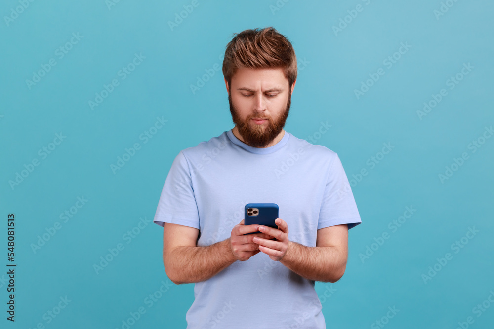 Portrait of serious concentrated bearded man texting message in social media on cell phone, using mobile network services, chatting online. Indoor studio shot isolated on blue background. Portrait of serious concentrated bearded man texting message in social media on cell phone, using mobile network services, chatting online. Indoor studio shot isolated on blue background.