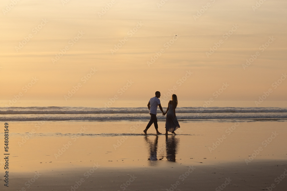 Loving couple walking on beach at sunset. Man and woman in casual clothes strolling along water at dusk. Love, family, nature concept