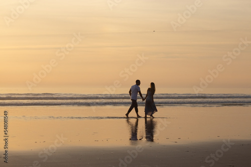 Loving couple walking on beach at sunset. Man and woman in casual clothes strolling along water at dusk. Love, family, nature concept