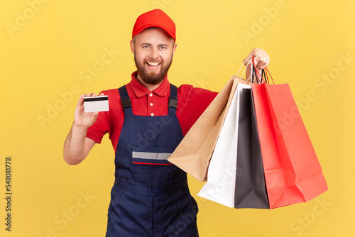 Portrait of positive smiling deliveryman in blue uniform, red t-shirt and hat holding shopping bag and credit card, cashless payments. Indoor studio shot isolated on yellow background.