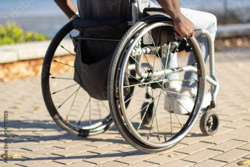 Strong Black man sitting in wheelchair outside enjoying his ride along wide grey tile road in city park on warm day. Sea background. Closeup shot. Disability, leisure time, motivation concept.
