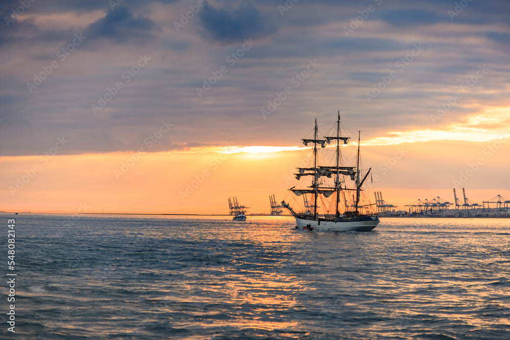 HONFLEUR, NORMANDY, FRANCE: Armada 2019 Grande Parade, tall ship Le Français sailing away in the Seine Estuary, in front of Le Havre harbour, with setting sun reflection in the sea