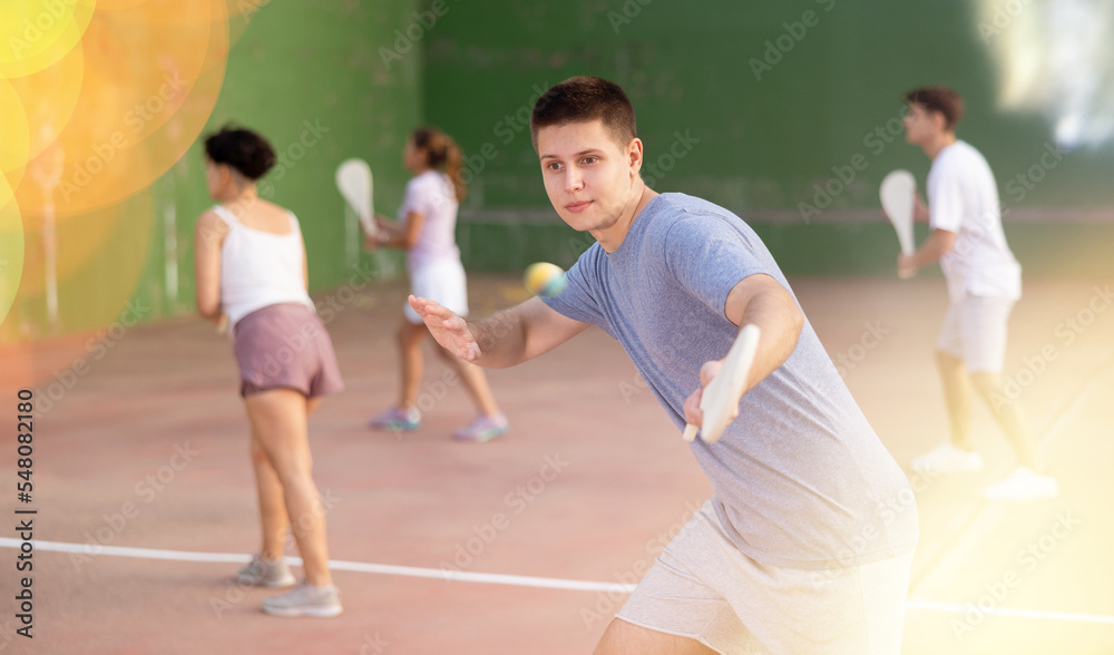 Portrait of emotional determined young guy playing pelota at open-air fronton in summer, swinging traditional wooden bat to return ball. Sportsman ready to hit volley