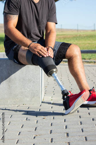 Close-up of male body with prosthetic leg. Trained guy with artificial leg sitting outdoors fixing his prothesis for street workout. Active life of people with disability, sport motivation concept