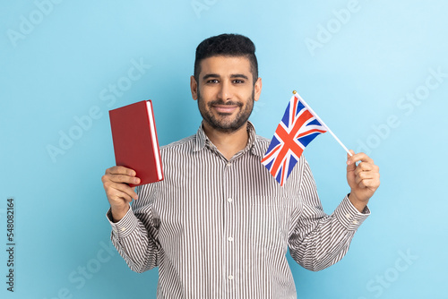 Portrait of attractive young adult man holding book and British flag, education abroad, looking at camera, wearing striped shirt. Indoor studio shot isolated on blue background. photo