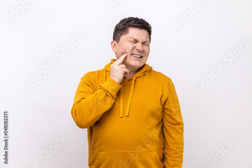 Unhealthy middle aged man wincing in pain and touching sore cheek, suffering unbearable toothache, gum disease, wearing urban style hoodie. Indoor studio shot isolated on white background.