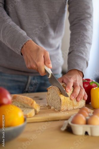 Close-up of mans hands cutting bread for healthy breakfast. Young man holding loaf of bread on wooden board preparing tasty sandwiches before going to work. Cooking as care of family health concept