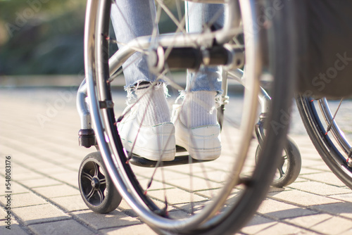 Person of unrecognizable gender and age sitting in wheelchair outdoors on sunny day. Close-up shot of thin legs in white sneakers and light blue jeans. Disability concept.