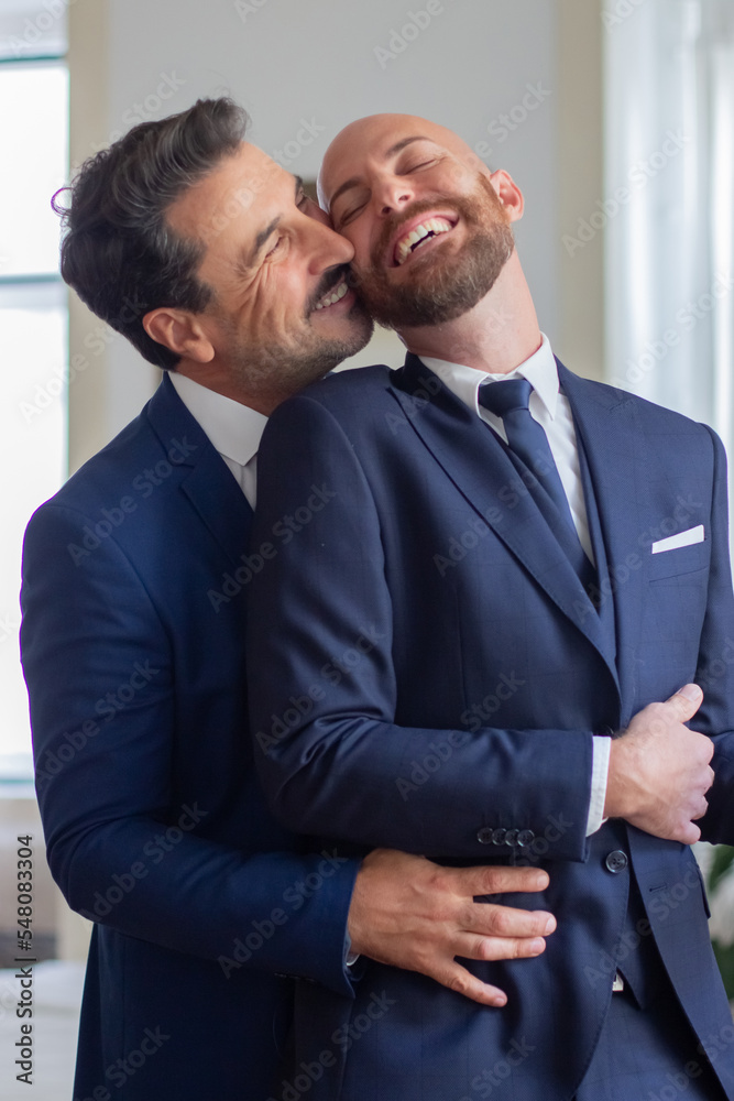 Happy gay in official suits hugging and kissing in hotel room. Vertical shot of handsome homosexual couple laughing, getting dressed for wedding ceremony, having fun together. LGBT, love concept
