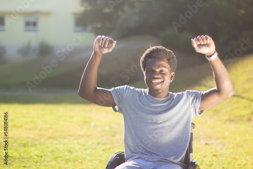 Portrait of young Black man in wheelchair enjoying sunny weather outdoors on green sunlit meadow. Cheerful guy with disability raising his thumbs in joy. Disability, motivation, happiness concept.