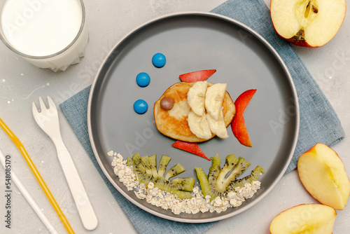 Funny fish face shape snack from pancake banana apples on plate. Cute kids childrens baby s breakfast sweet dessert  healthy lunch  food art with milk kiwi on gray concrete background top view