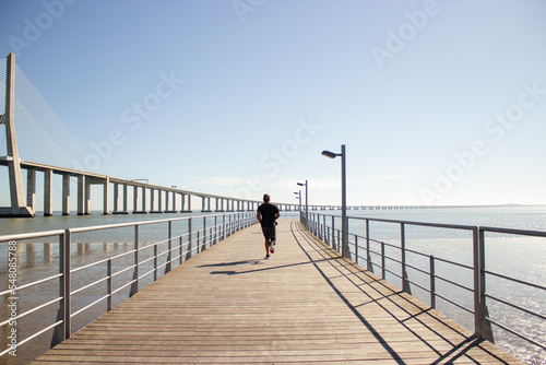 Sporty man with disability enjoying sport exercises in summer. Young man in T-shirt and shorts training in summer running along high bridge over sea. Sport practice of people with disability concept