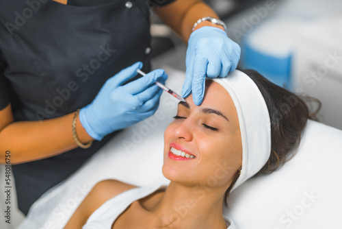 Eyebrow filler injection concept. Closeup indoor portrait of smiling caucasian woman with her head placed on SPA bed during collagen injection performed by professional expert in protective gloves