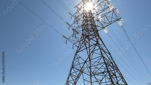 Large electrical tower and power lines against blue sky and bright sun, tilt up; in Ontario Canada.