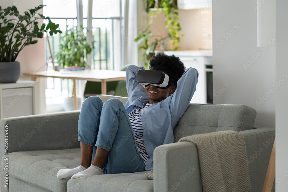 Young joyful African woman wearing modern VR headset sitting on sofa in living room, watching immersive 3D video, overjoyed black girl using virtual reality glasses at home. Future technologies Young joyful African woman wearing modern VR headset sitting on sofa in living room, watching immersive 3D video, overjoyed black girl using virtual reality glasses at home. Future technologies
