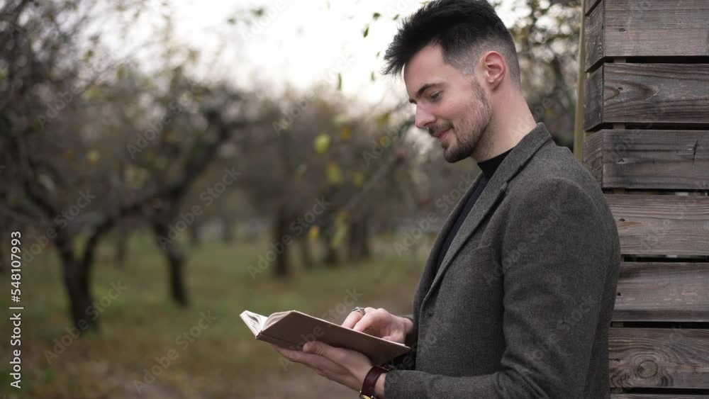 Side view intelligent confident handsome man reading book standing in autumn garden laughing. Positive Caucasian guy in suit enjoying hobby outdoors on overcast day. Lifestyle and leisure Side view intelligent confident handsome man reading book standing in autumn garden laughing. Positive Caucasian guy in suit enjoying hobby outdoors on overcast day. Lifestyle and leisure