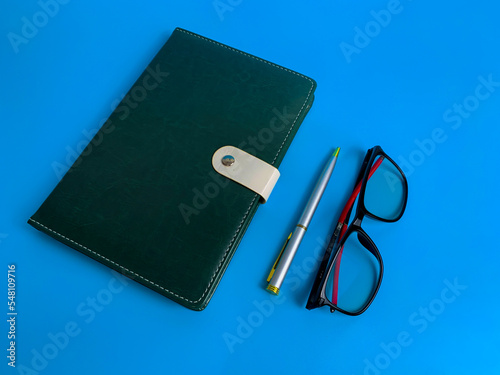 top view blank green leather diary, pen and glass on blue background