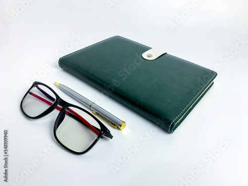 Closeup blank green leather diary, pen and glasses on white desk