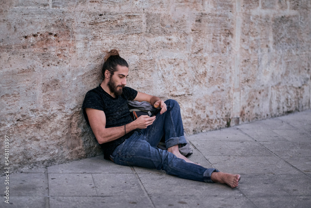 Caucasian young man with long hair barefoot calm and relaxed sitting on the floor next to a large backpack leaning against a stone wall looking at the screen of his smartphone