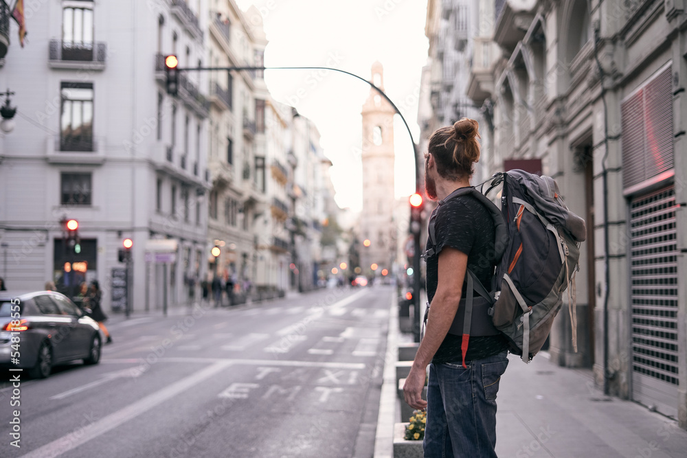 young caucasian man with a beard and long hair standing on a city street with a backpack on his back, valencia, spain