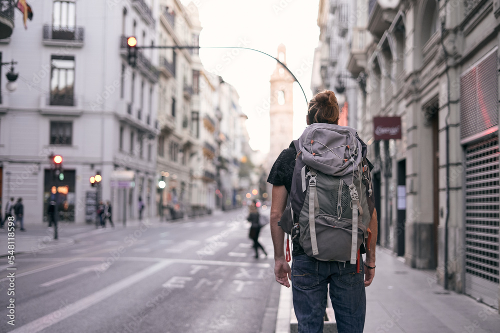 young caucasian man standing with his back on a city street with a large backpack on his back, valencia, spain young caucasian man standing with his back on a city street with a large backpack on his back, valencia, spain