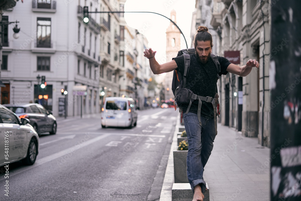 caucasian guy with a beard and long hair with a backpack on his back barefoot jumping on the city street, valencia, spain