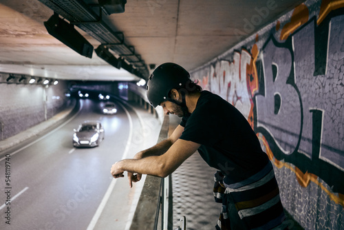 young caucasian man standing with helmet on his head leaning on the railing of a walkway inside a tunnel with a lot of traffic in the city