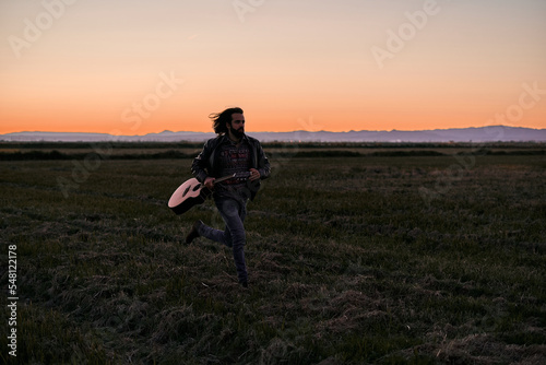 Caucasian young man with long hair in fur jacket running across the plain away from the mountains holding a guitar at sunset