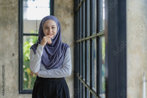 Confident beautiful muslim businesswoman standing with folded arms near window working in office, modern muslim businesswoman portrait