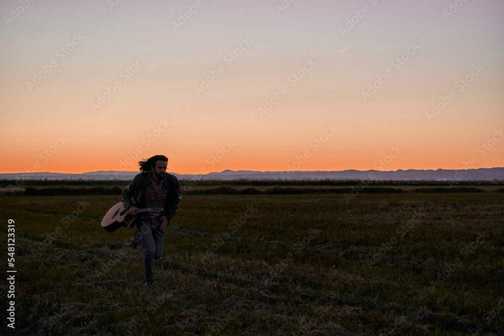 caucasian young man in fur jacket running across the big lonely green field next to his guitar at sunset
