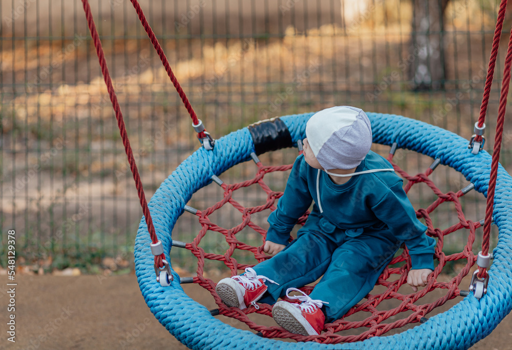 Cute Little boy with down syndrome in a funny hat walks in the playground, swinging on a swing