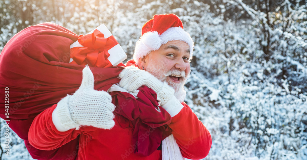 Obraz premium Santa claus with bag full of christmas presents outdoor. Santa portrait close up with thumbs up on winter snowy background. Santa Claus pulling huge bag of gifts on outside.