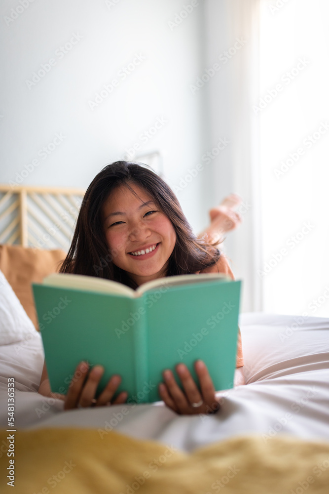 Vertical portrait of happy teen asian college student reading book at home lying on bed in cozy room looking at camera. Vertical portrait of happy teen asian college student reading book at home lying on bed in cozy room looking at camera.