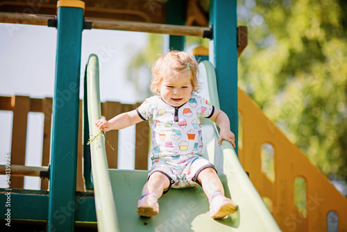 Happy blond little toddler girl having fun and sliding on outdoor playground. Positive funny baby child smiling. Summer leisure for small children, active games for family outdoors.