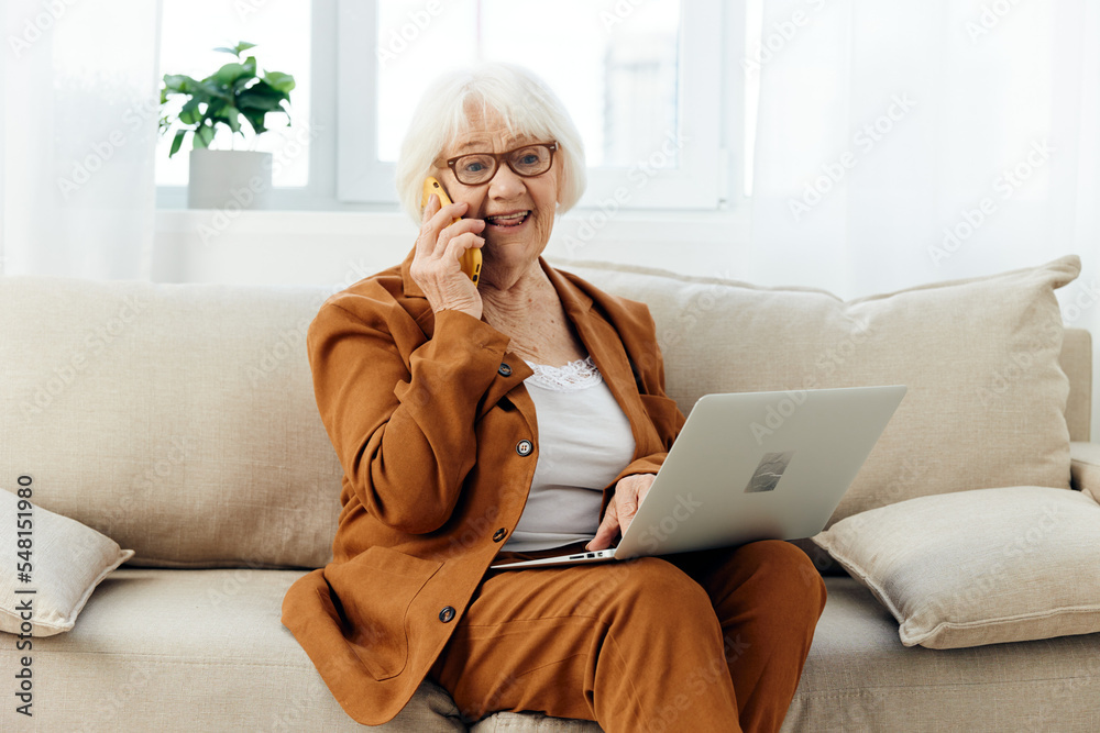 a happy elderly woman smiles sweetly sitting on the sofa in a bright apartment near the window and talking on a smartphone holding a laptop on her lap a happy elderly woman smiles sweetly sitting on the sofa in a bright apartment near the window and talking on a smartphone holding a laptop on her lap