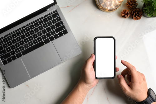 Top view of man hand using smartphone and laptop computer, lightbulb, pine cone on marble table