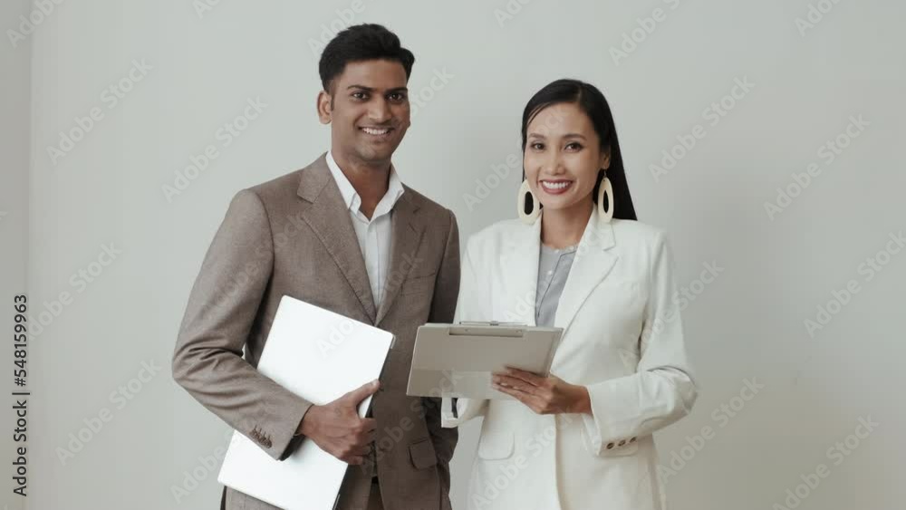 Medium shot of Asian businesswoman and Indian businessman reading something on clipboard and then posing for camera and smiling Medium shot of Asian businesswoman and Indian businessman reading something on clipboard and then posing for camera and smiling