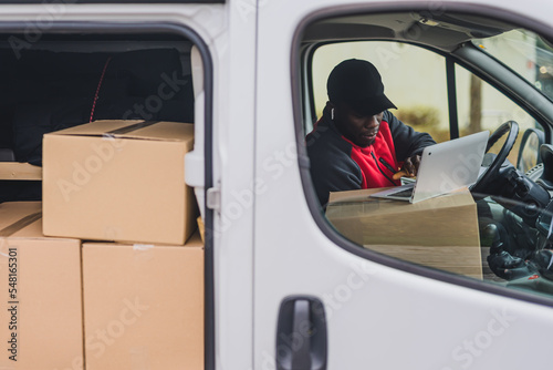 Outdoor perspective on focused hard-working African-American 30-year-old man working on his laptop in parcel van. Side door of white delivery van showing many cardboard packages ready to be delivered