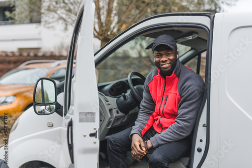 Occupation - delivery person. Positive smiling Black 25-year-old man dressed in postal delivery uniform in red and black color sitting on driver's seat. High quality photo