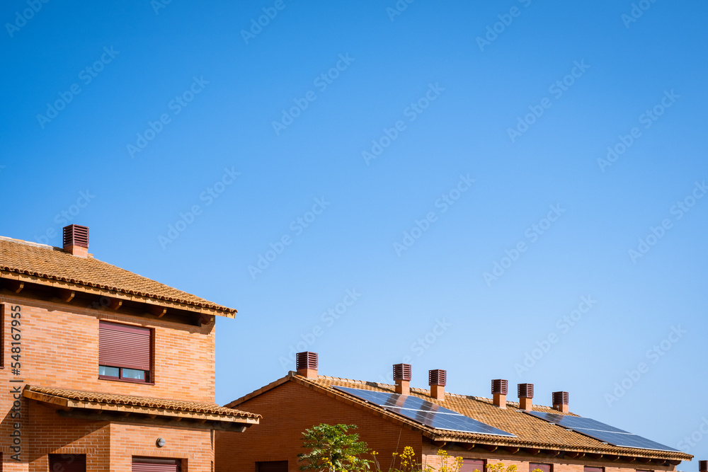 Single-family house, two-story, red adobe brick, with several black solar panel panels on its red tile roof. Clear blue sky, no clouds. Autumn.  Tenerife, Canary Islands, Spain.