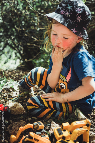 Small child resting and relaxing while sitting on a rock sucking on his fingers, taking a break while playing with diggers in a natural park. Overcoming difficulty and self soothing.