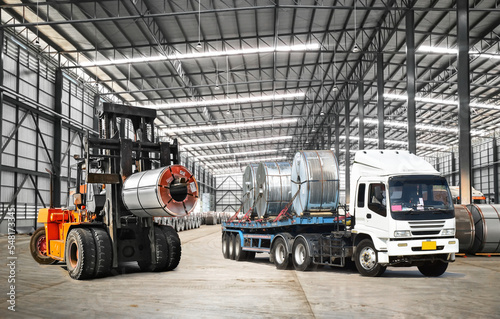 Forklift lifts heavy steel coil inside a warehouse at a seaport-bounded area to load onto truck. Raw material handling and transportation. © Hor