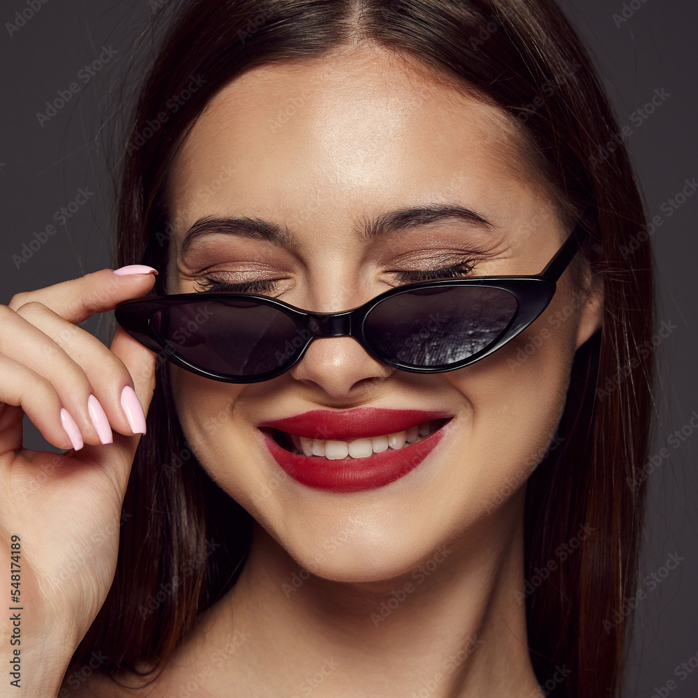Close-up portrait of young beautiful girl with red lips posing in sunglasses, smiling isolated over dark grey background. Concept of beauty, fashion and cosmetology Close-up portrait of young beautiful girl with red lips posing in sunglasses, smiling isolated over dark grey background. Concept of beauty, fashion and cosmetology