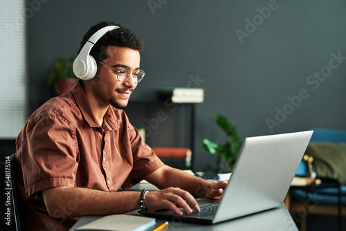 Young smiling man in headphones typing on laptop keyboard while sitting by workplace and taking part in online webinar or lesson photo